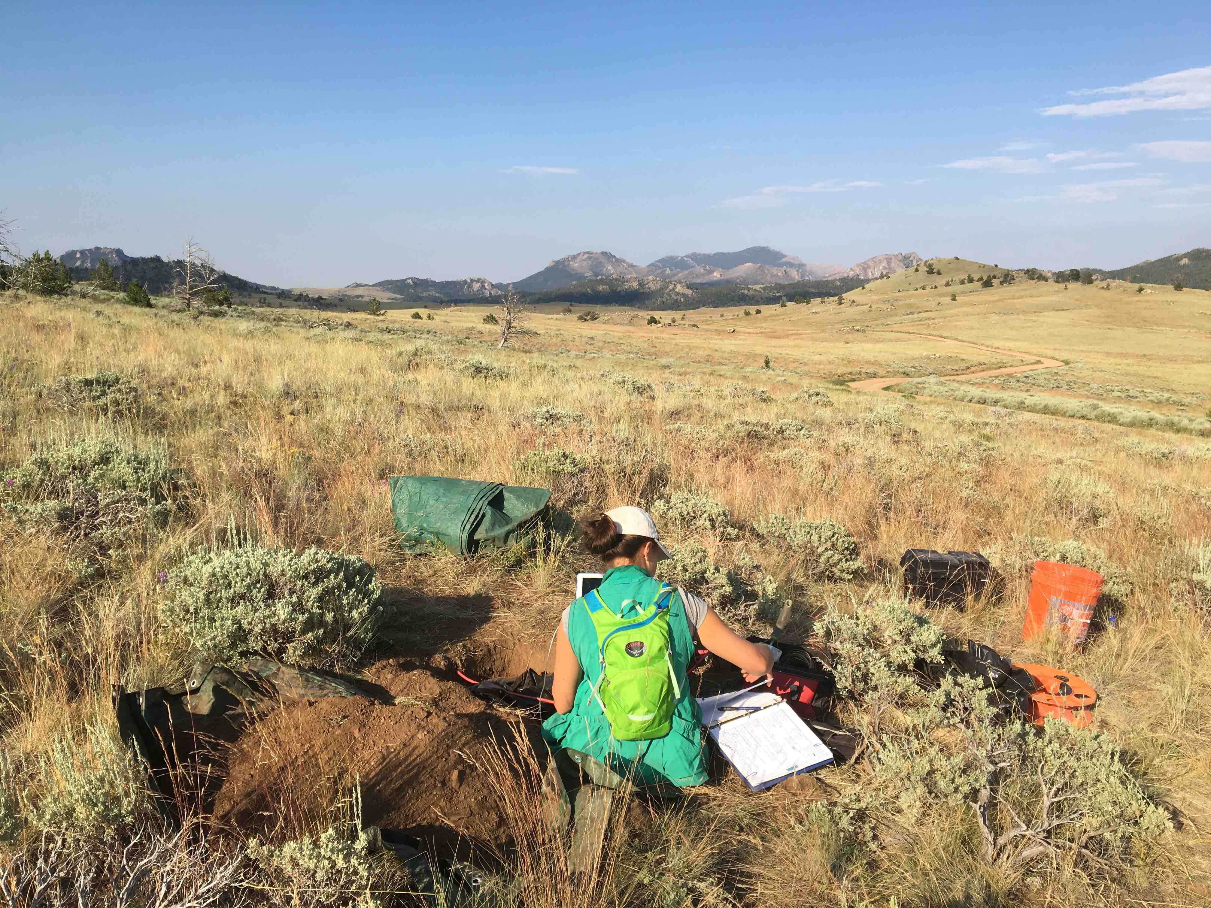 Eva sitting in a dry grassy field with a view of mountains in the background. Starting magnetotelluric data acquisition, Wyoming.